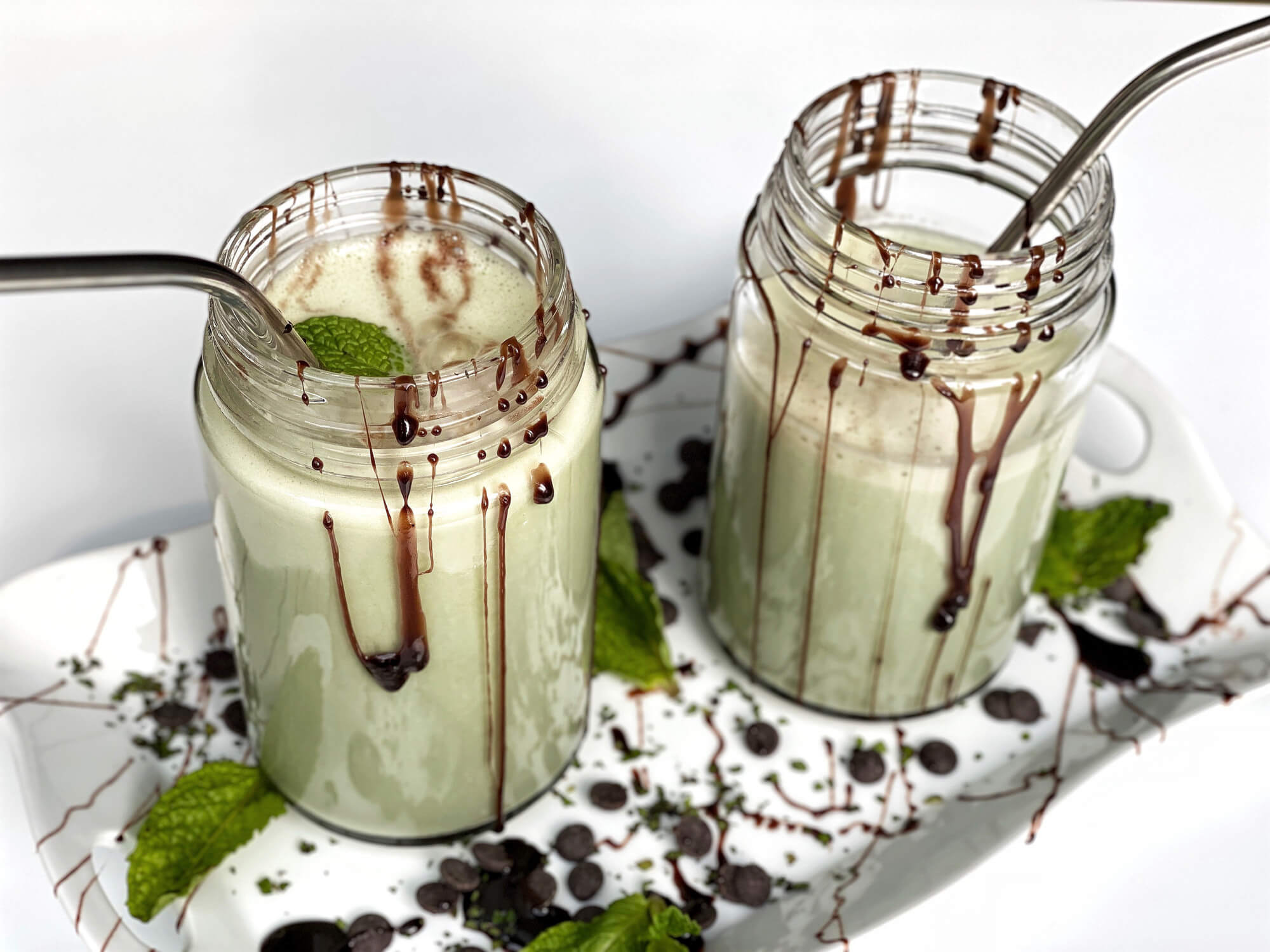 two chocolate mint shakes with straws in mason jars on a decorative tray dribbled in chocolate with mint leaves. white background.