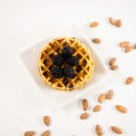 Top view - Blackberry Almond Chaffle on a white plate, topped with blackberries and surrounded by whole almonds.