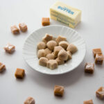Cream colored Fat Bombs on a white plate, surrounded by caramels and a stick of butter.