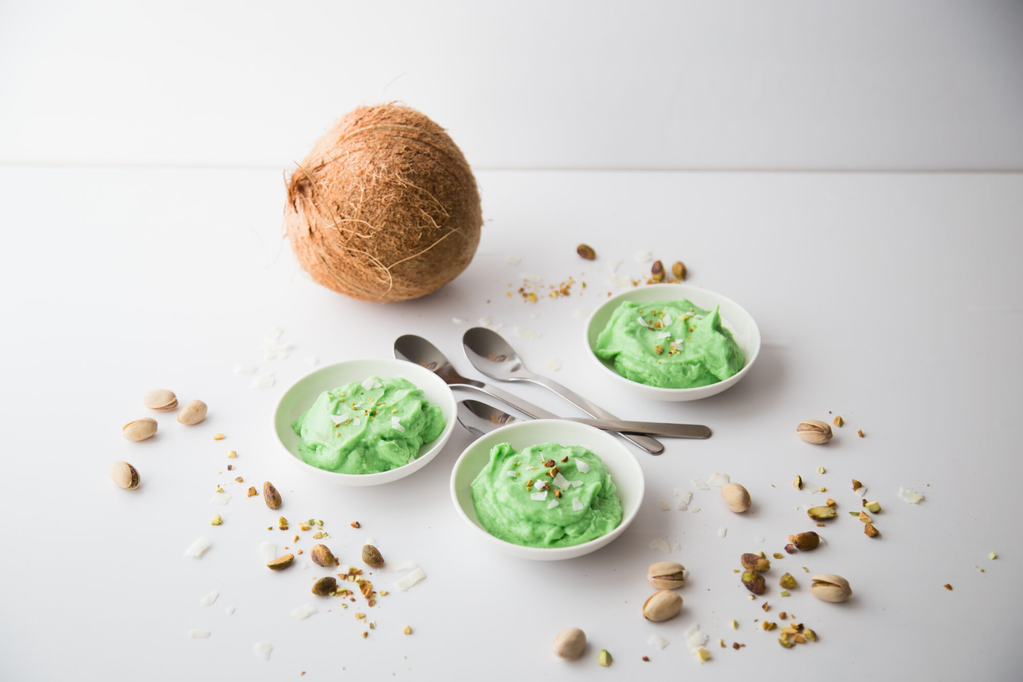 Three bowls filled with green pistachio pudding, topped with crushed pistachios, surrounded by three spoons, a coconut and more crushed pistachios.