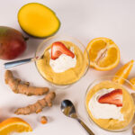 Top view - Two short mousse cups filled with Mango Mousse, topped with cream and a strawberry. Desserts are surrounded by ginger, spices and mangoes.