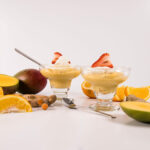 Two short mousse cups filled with Mango Mousse, topped with cream and a strawberry. Desserts are surrounded by ginger, spices and mangoes.