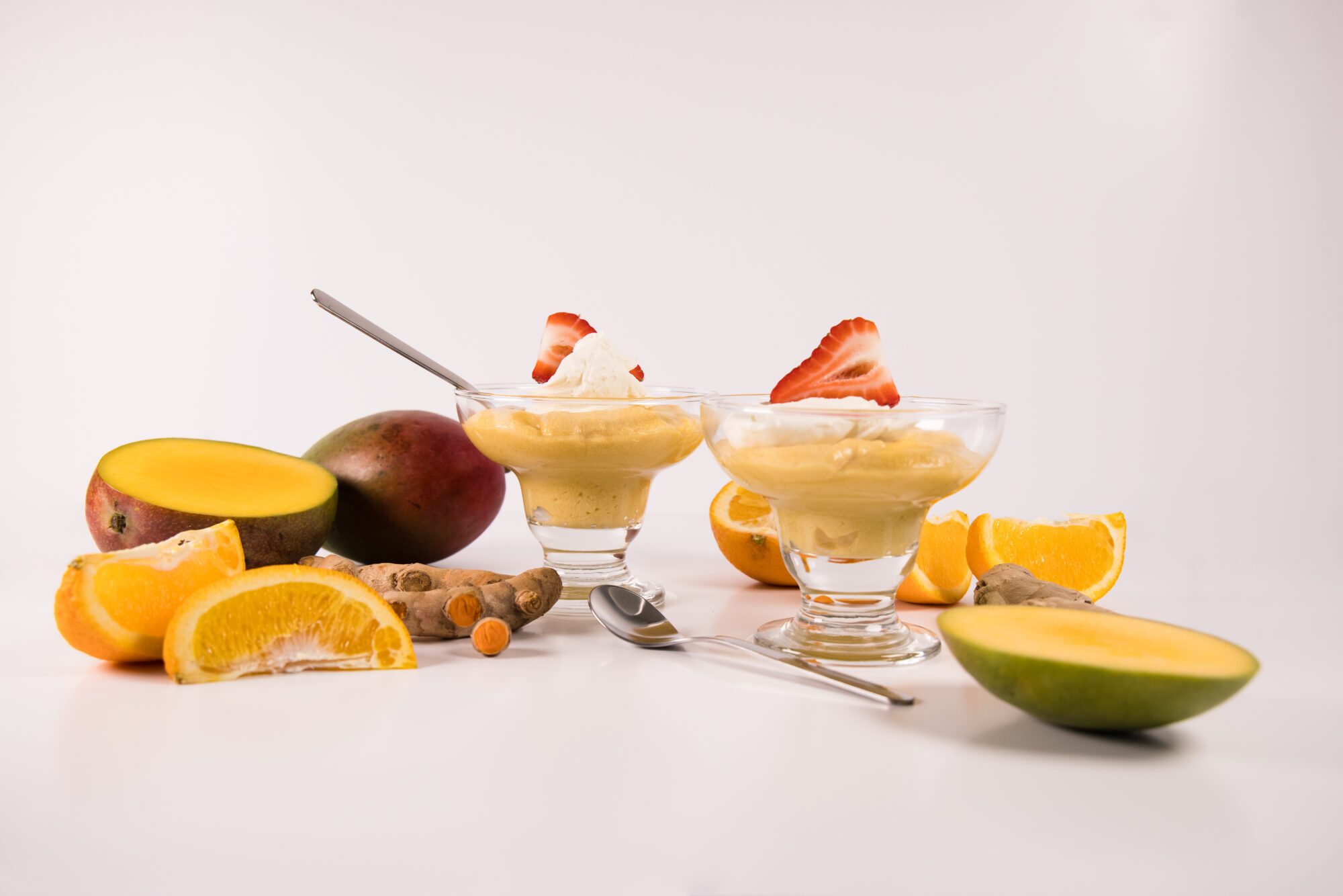 Two short mousse cups filled with Mango Mousse, topped with cream and a strawberry. Desserts are surrounded by ginger, spices and mangoes.