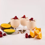 Three glass dessert cups filled with Mango Orange Yogurt, topped with almonds and raspberries. Raspberries and sliced oranges and mangoes surround the desserts.