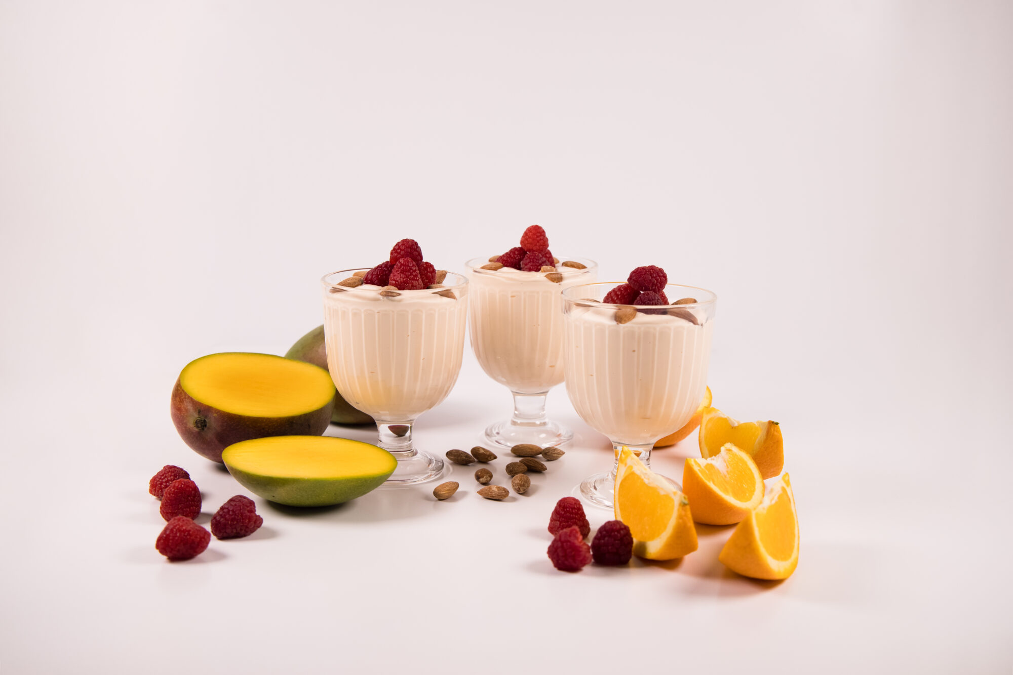 Three glass dessert cups filled with Mango Orange Yogurt, topped with almonds and raspberries. Raspberries and sliced oranges and mangoes surround the desserts.