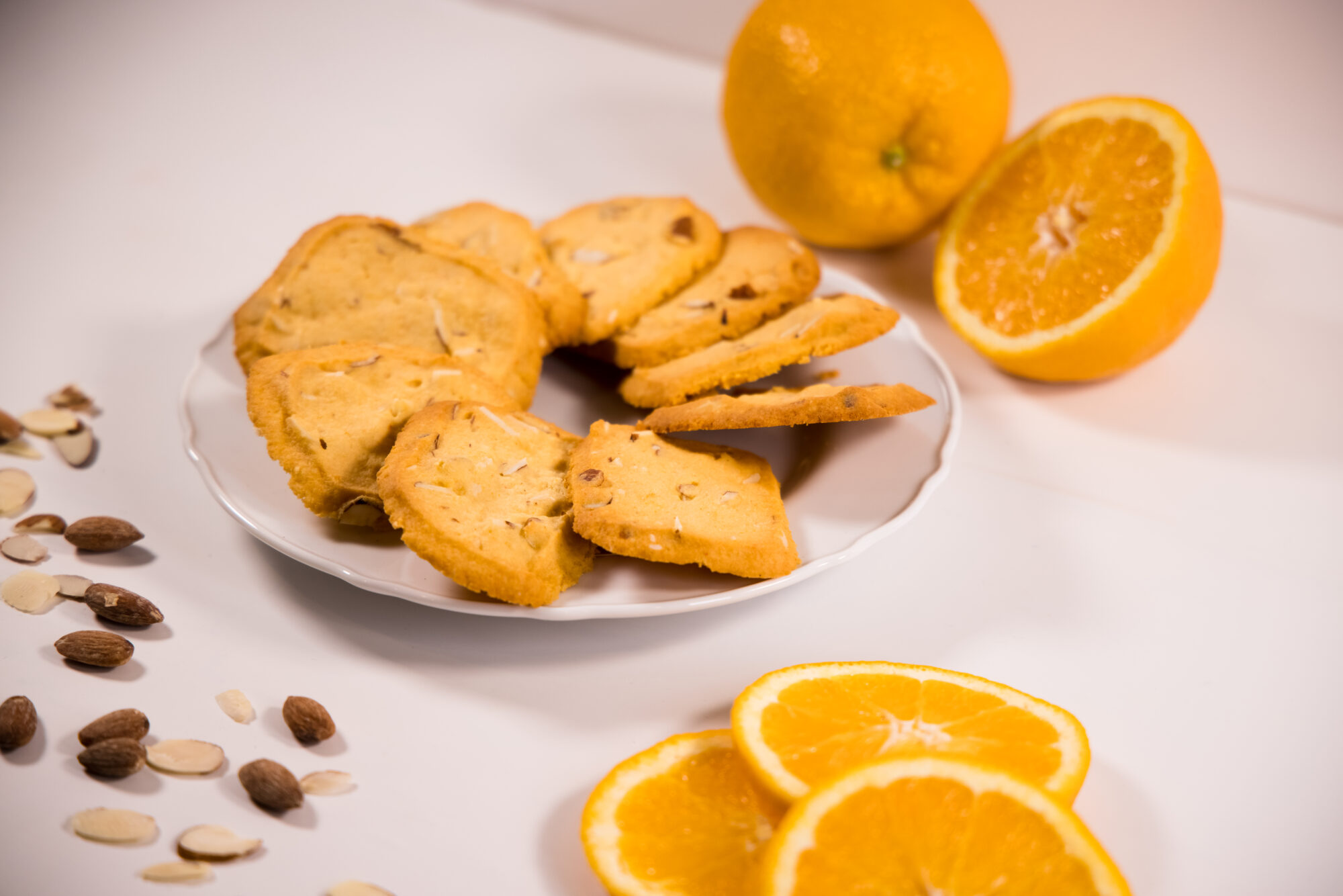 A white plate arranged with Orange Almond Shortbread cookies, surrounded by sliced oranges and almonds.
