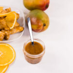 Close up - Diced chicken covered in orange sauce on a white plate. Sliced oranges, mangoes, and a jar of orange sauce to the side.