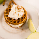 White plate with two chaffles and ice cream in the middle. Topped with whipped cream and sliced green apples on the side.