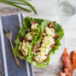 Top view of egg salad on top of bacon slices, lying on two lettuce leaves. A towel, fork, and bacon strips to the side.