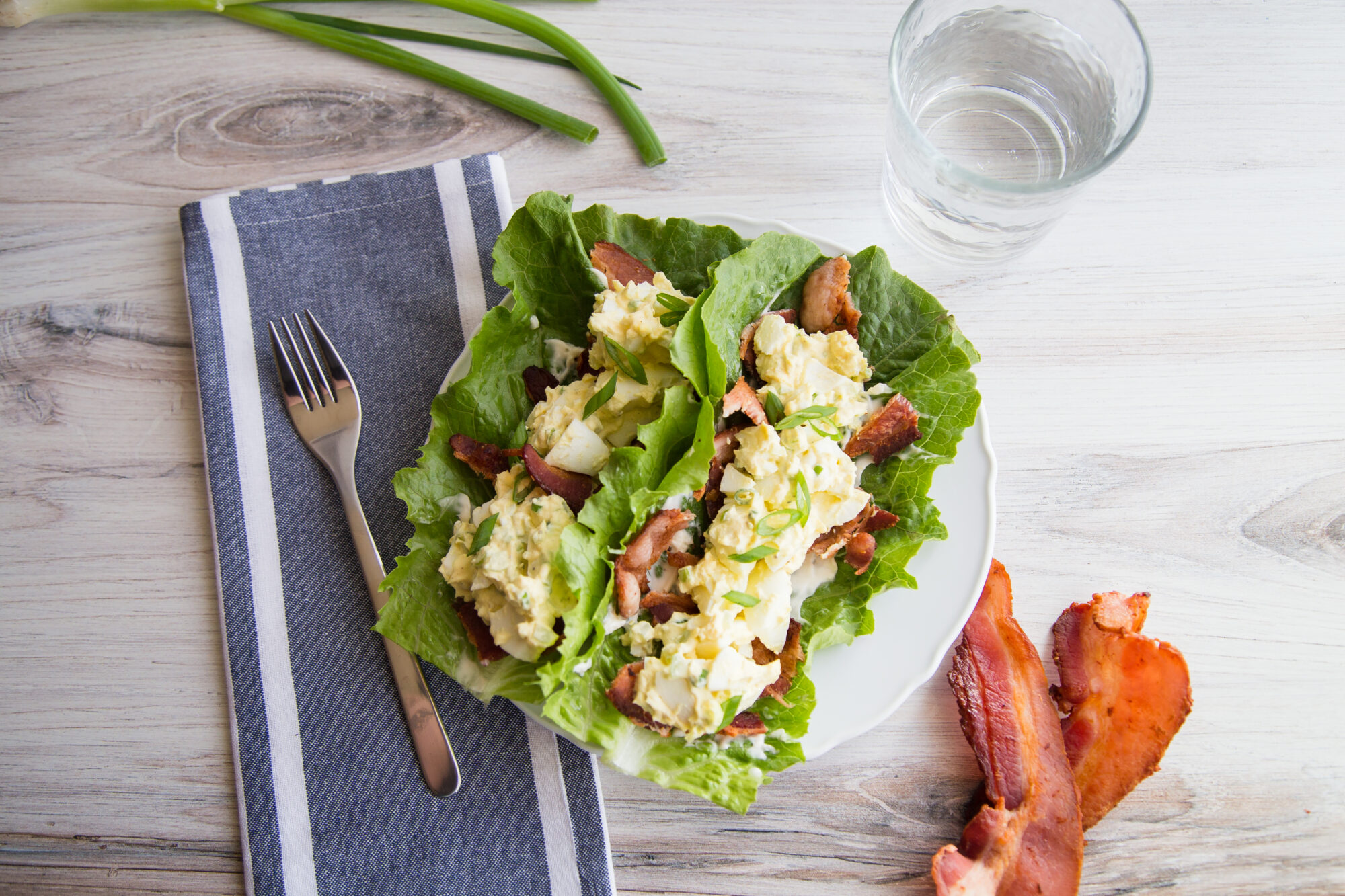 Top view of egg salad on top of bacon slices, lying on two lettuce leaves. A towel, fork, and bacon strips to the side.