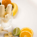 Top view - Three Banana Daquiri popsicles in a glass jar with two more laying to the side. Slices of bananas, oranges and limes surround the jar.