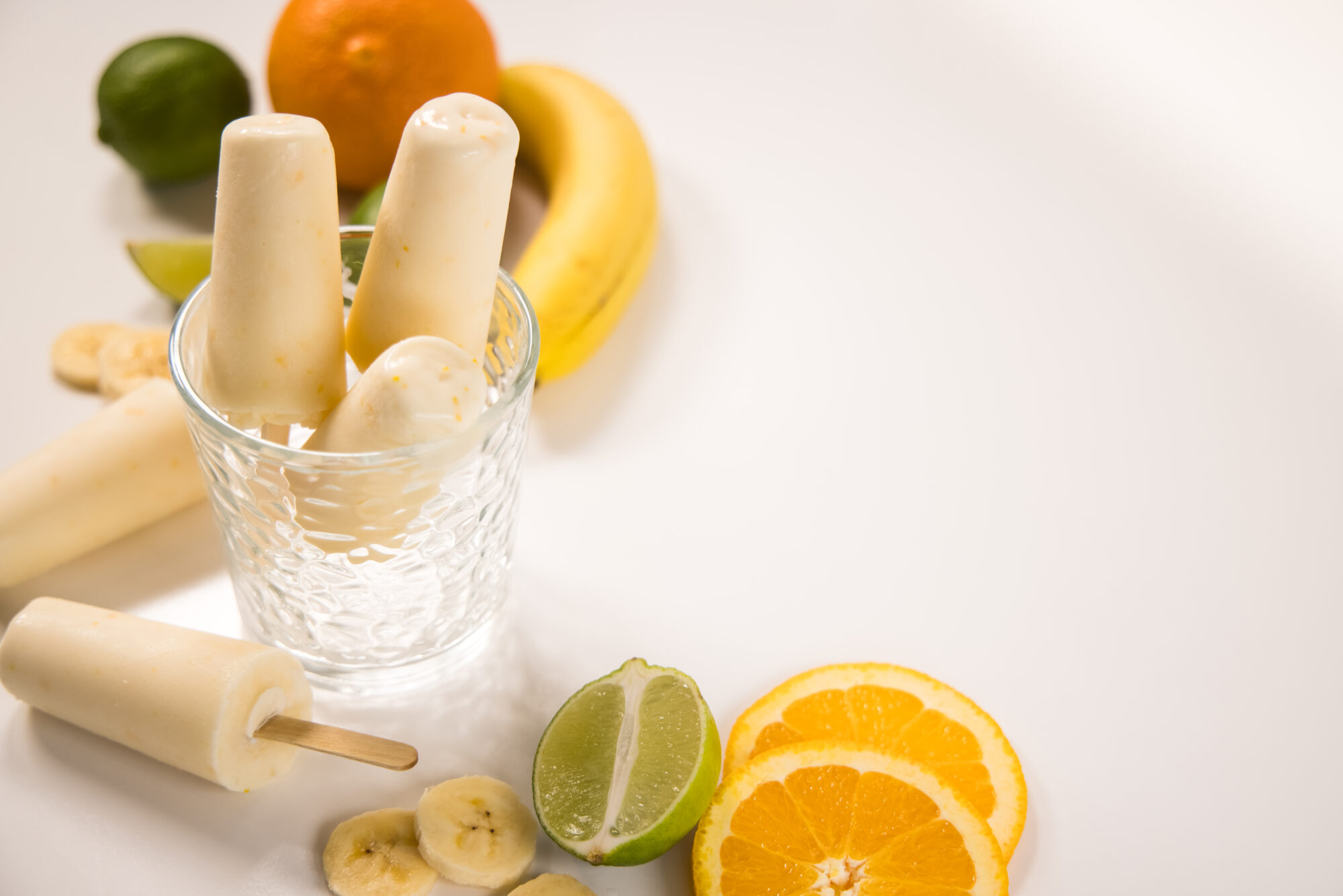 Top view - Three Banana Daquiri popsicles in a glass jar with two more laying to the side. Slices of bananas, oranges and limes surround the jar.