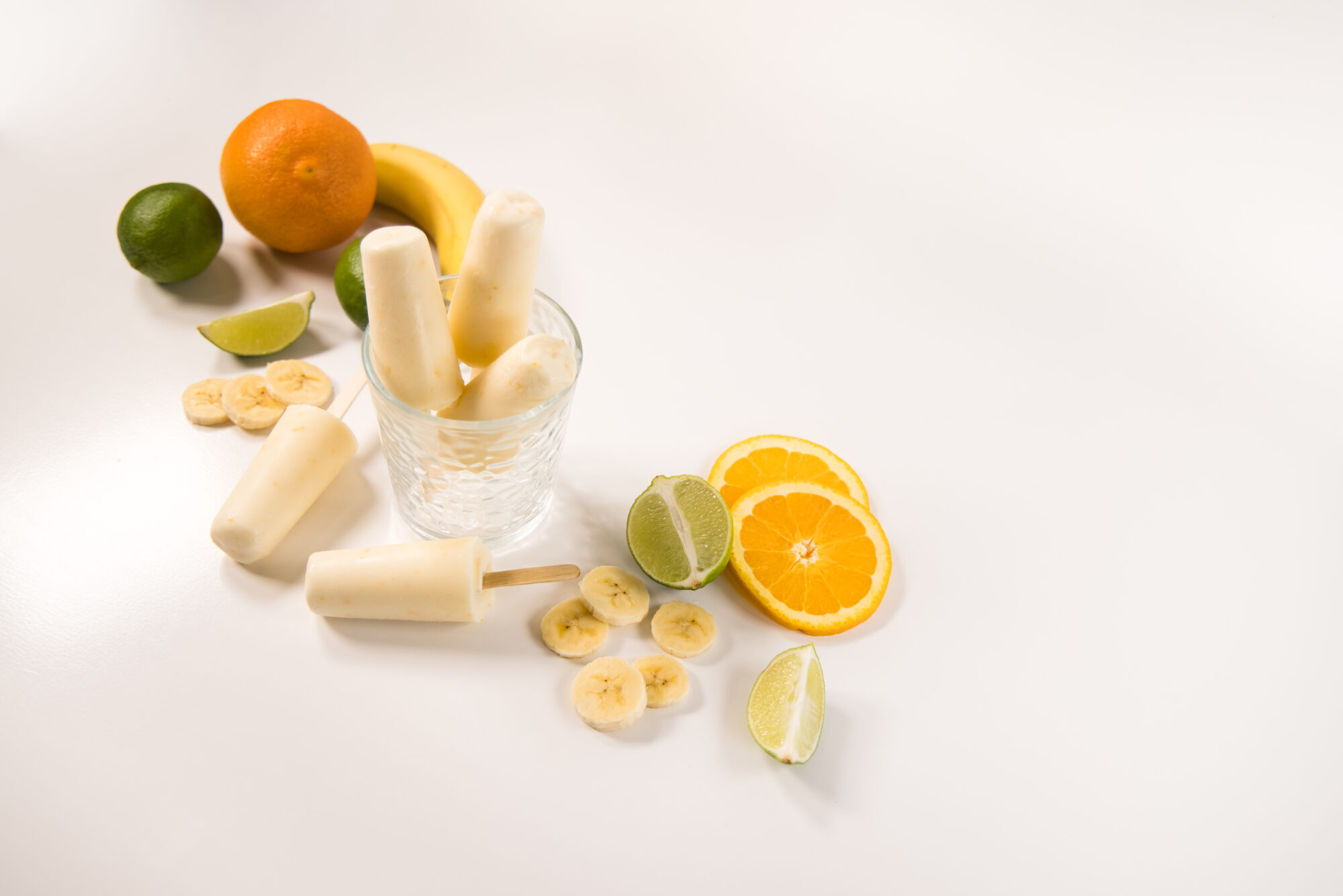 Three Banana Daquiri popsicles in a glass jar with two more laying to the side. Slices of bananas, oranges and limes surround the jar.