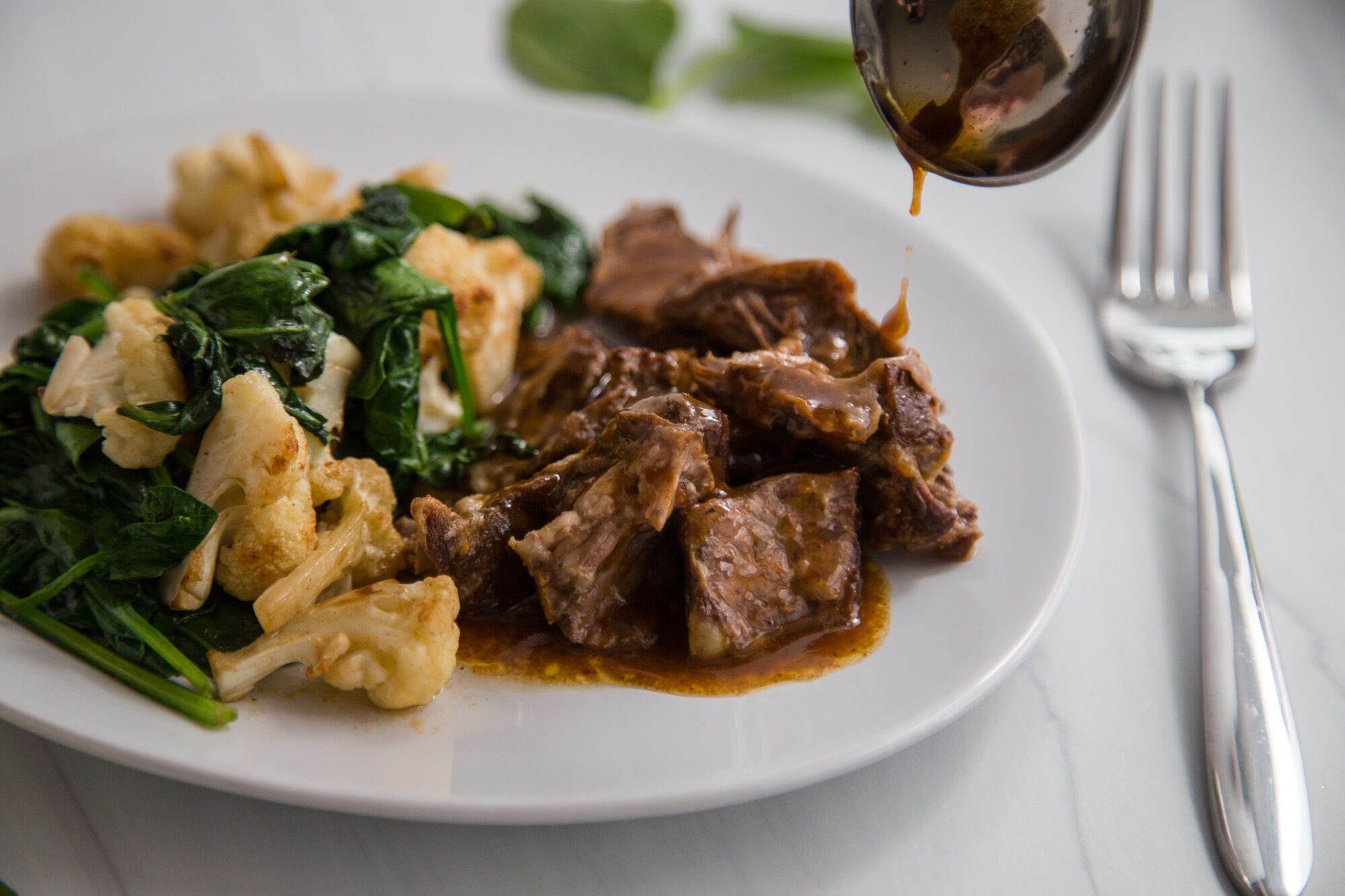Close up- white plate with braised chuck roast with gravy, cauliflower and cooked spinach.