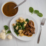 Top view-white plate with braised chuck roast in gravy, cauliflower and cooked spinach. On the table; a cup with gravy, spinach leaves and a fork.
