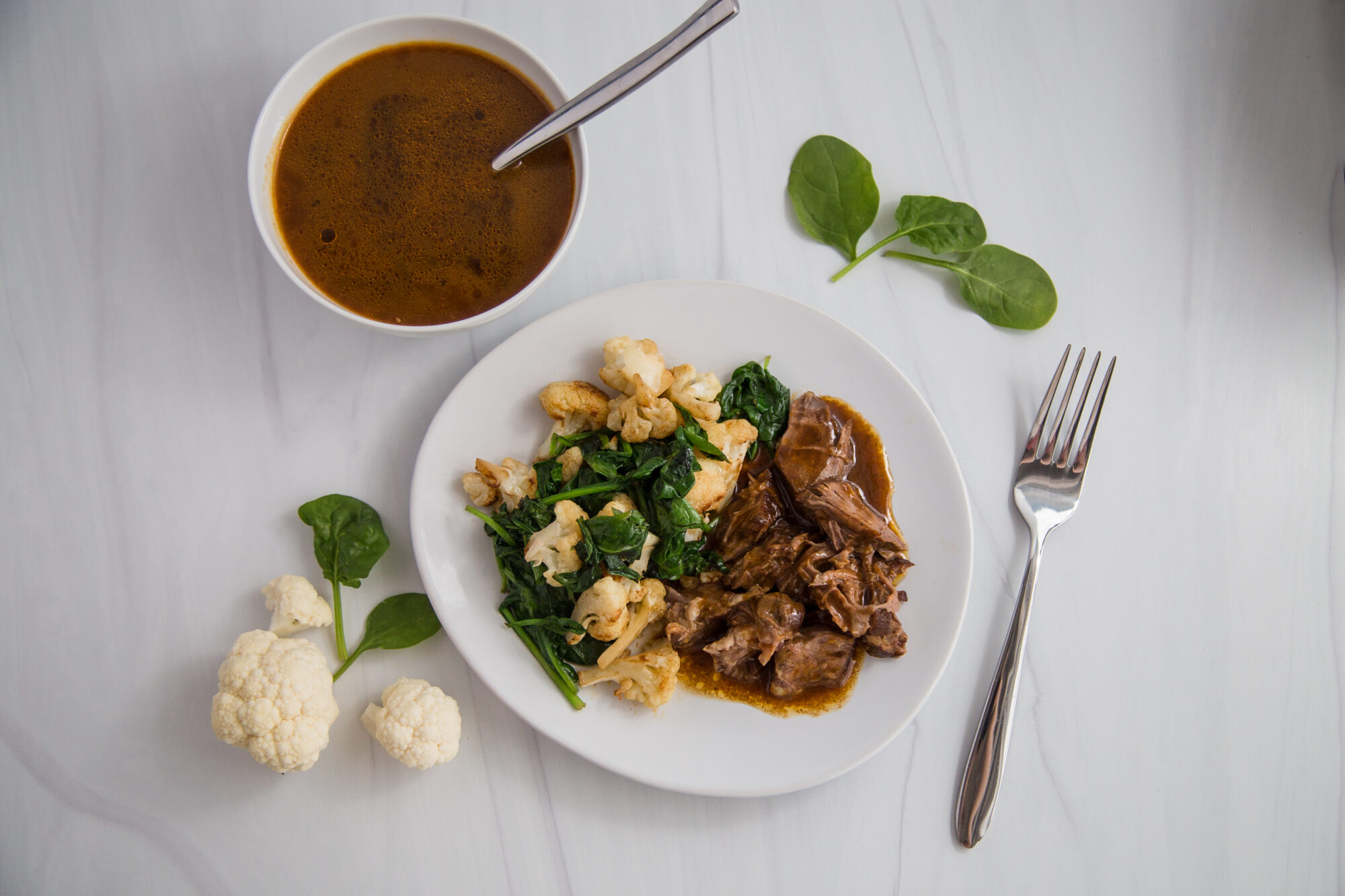 Top view-white plate with braised chuck roast in gravy, cauliflower and cooked spinach. On the table; a cup with gravy, spinach leaves and a fork.