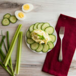 Top view-White plate, Egg salad on a half an avocado, surrounded by sliced cucumber. On the table is a red napkin, fork, celery, cucumber and a hard boiled egg, sliced in half