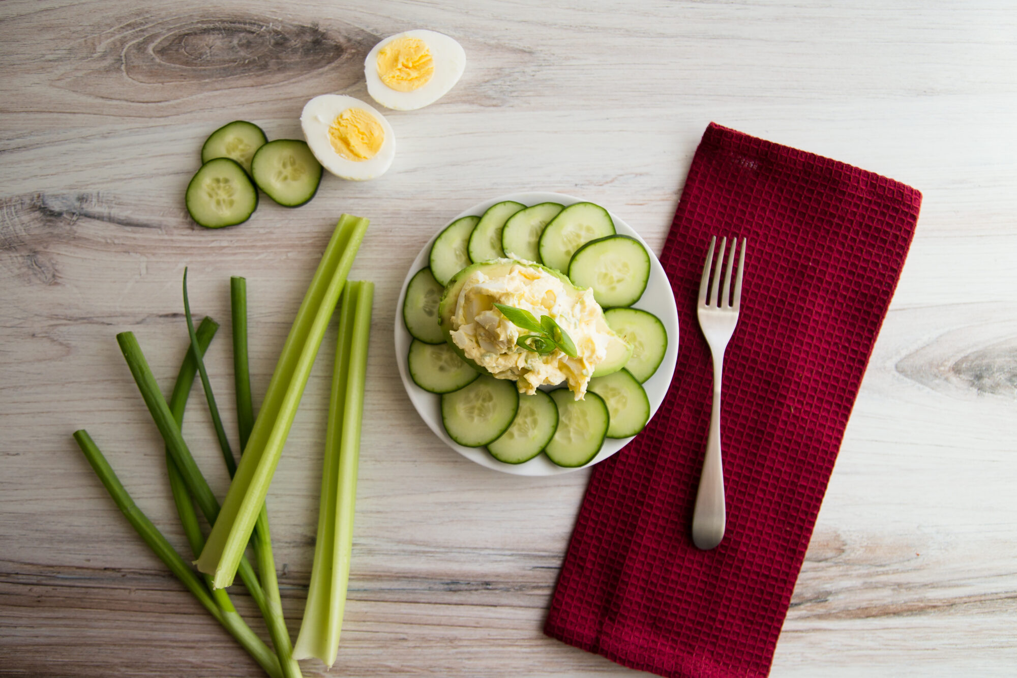 Top view-White plate, Egg salad on a half an avocado, surrounded by sliced cucumber. On the table is a red napkin, fork, celery, cucumber and a hard boiled egg, sliced in half
