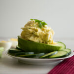 Close up view- Egg salad on a half an avocado, surrounded by sliced cucumber on a white plate