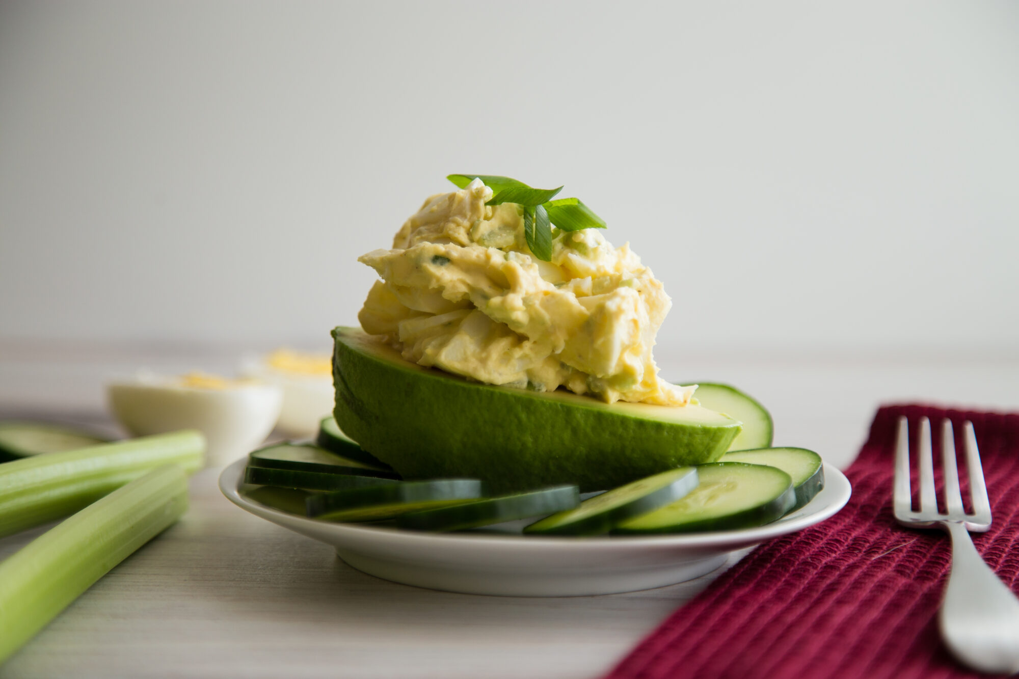 Close up view- Egg salad on a half an avocado, surrounded by sliced cucumber on a white plate