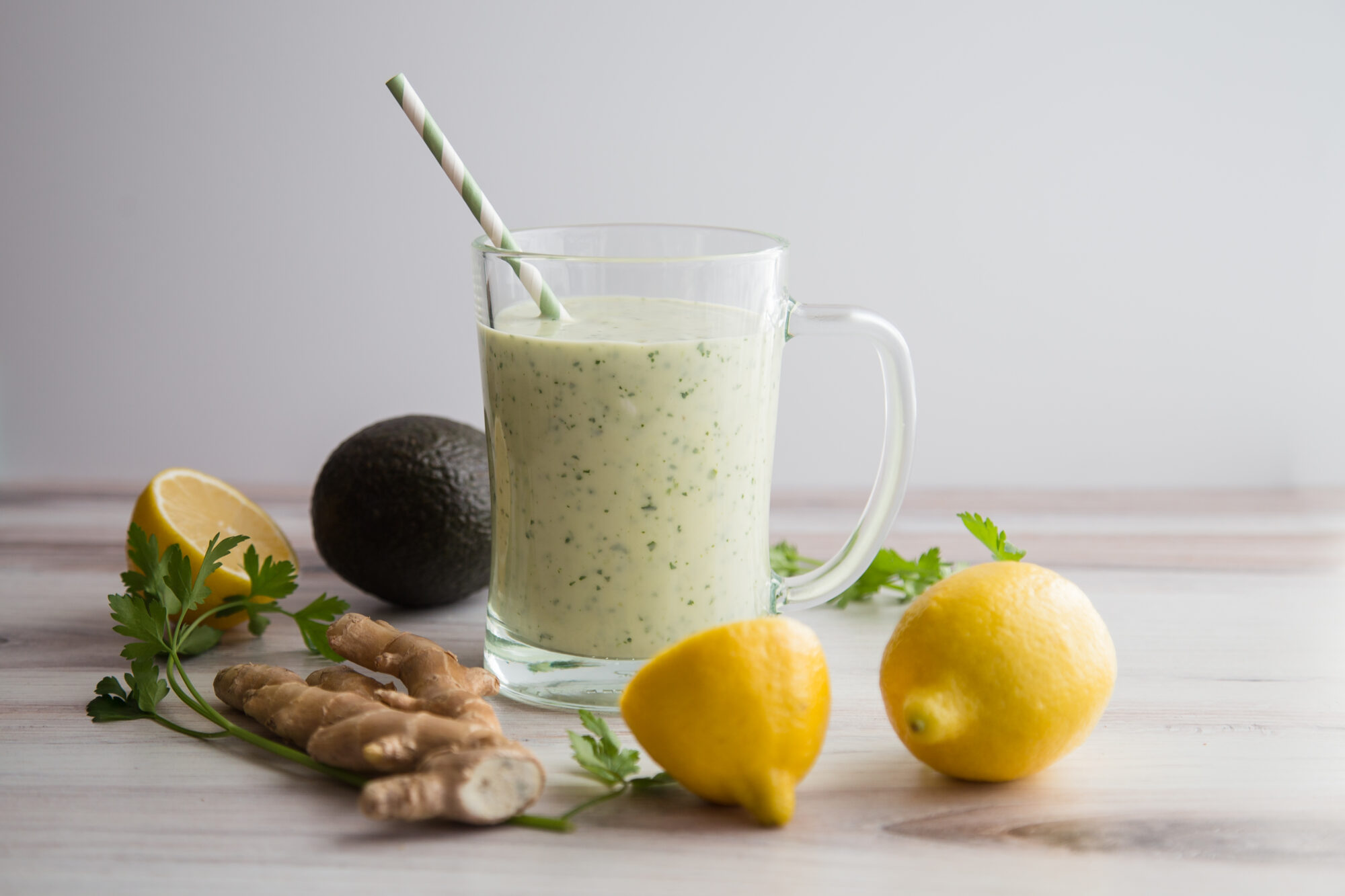 Distant view-Close up-glass mug with green smoothie. Green and white straw in the smoothie. Surrounded by lemons, ginger and a coconut.