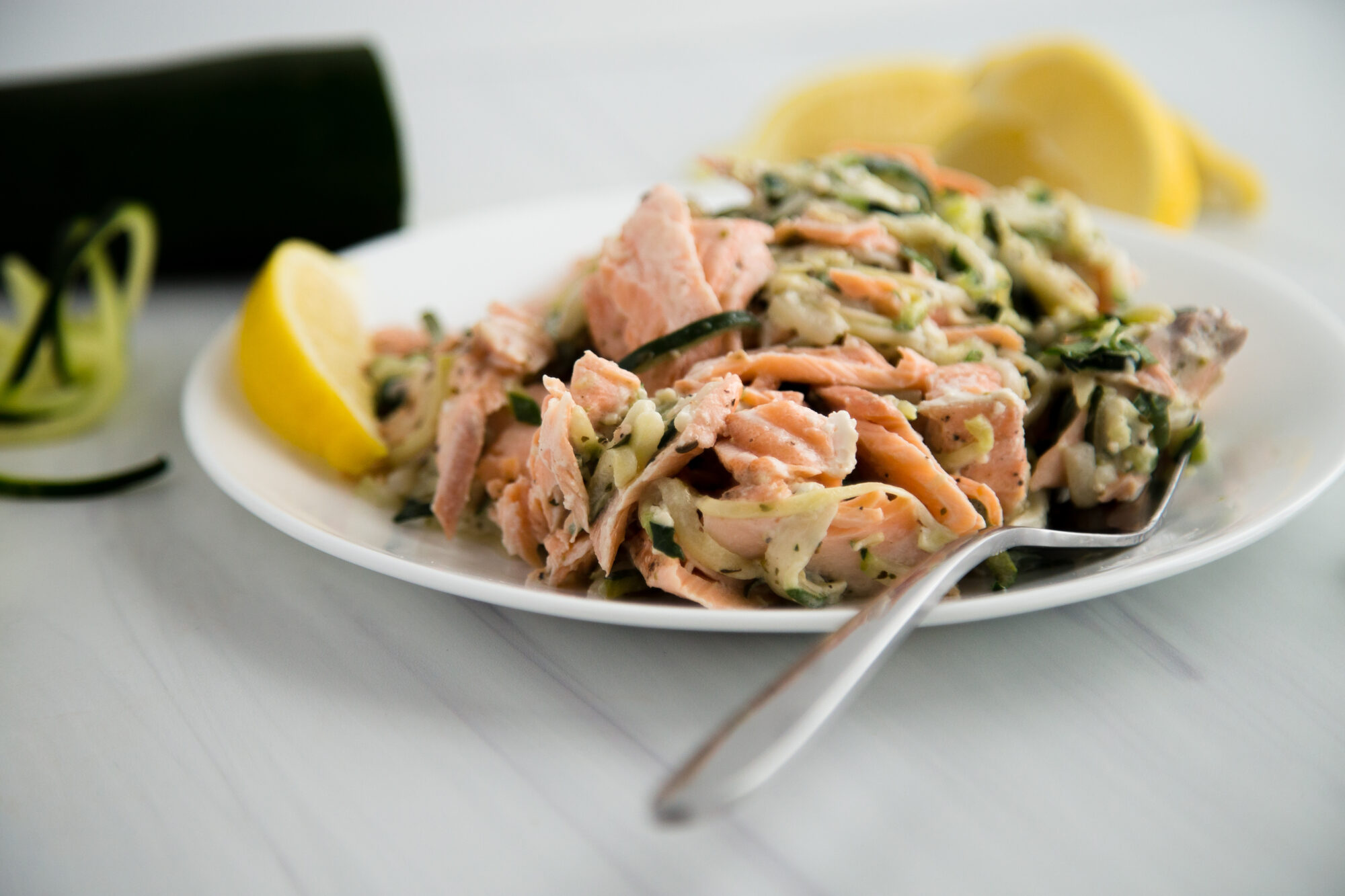 Close up of salmon and zucchini noodles on a white plate with a lemon wedge and a fork