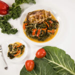 Close up - Two white plates with simmered collard greens, kale and red peppers. The larger plate also has a ham bone. Next to the plates are collard and kale leaves and roma tomatoes.