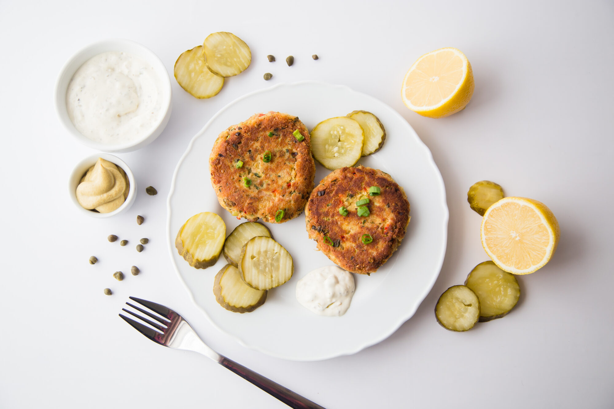Top view- Two tuna patties on a white plate with pickle chips and lemon wedges.