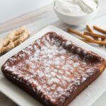 Square Ginger cake sprinkled with powdered erythritol, on a white plate. Ginger root, cinnamon sticks, and a bowl of Chantilly Cream sits to the side.
