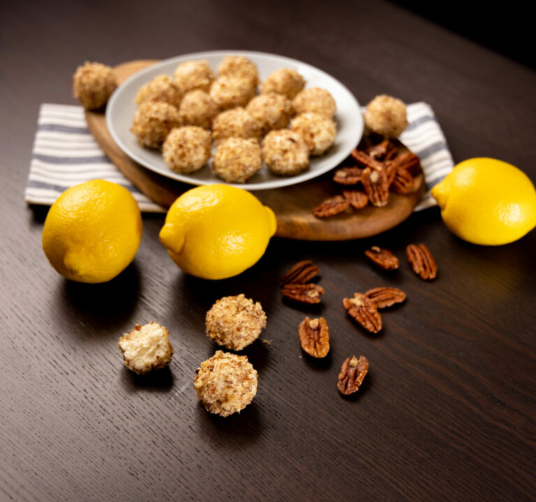 White plate filled with Lemon Cheesecake Bites. Surrounded by lemons and pecans.