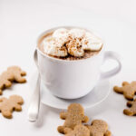 White mug filled with Hot Gingerbread Cocoa, topped with whipped cream. Gingerbread cookies to the side.