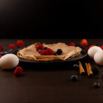 Close up - Sweet Cinnamon Egg White Protein Crepes, topped with berries. Eggs, cinnamon sticks and berries to the side.