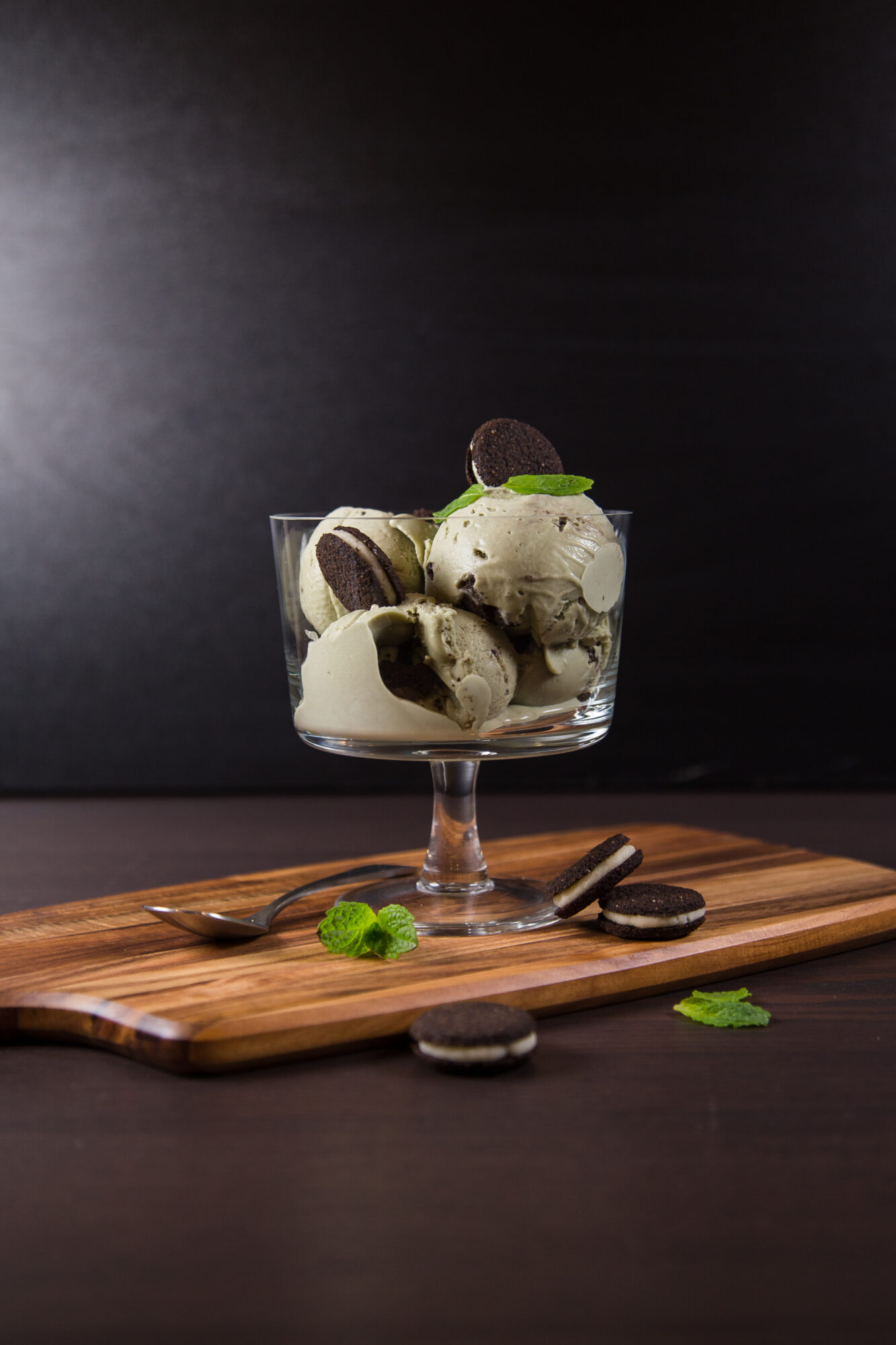 Side view - Wooden cutting board with a glass dish filled with Mint Chocolate Cookie Ice Cream, topped with cookies.
