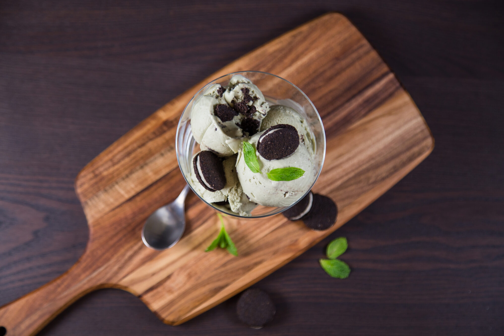 Wooden cutting board with a glass dish filled with Mint Chocolate Cookie Ice Cream, topped with cookies.