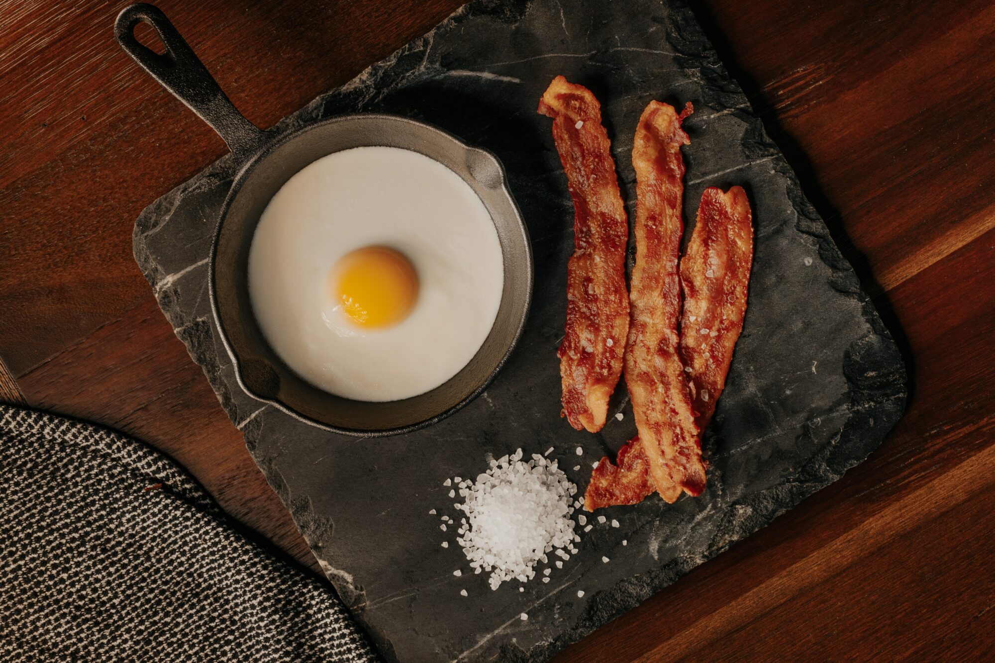 Cast iron skillet with an easy over egg. Three strips of cooked bacon on the side.