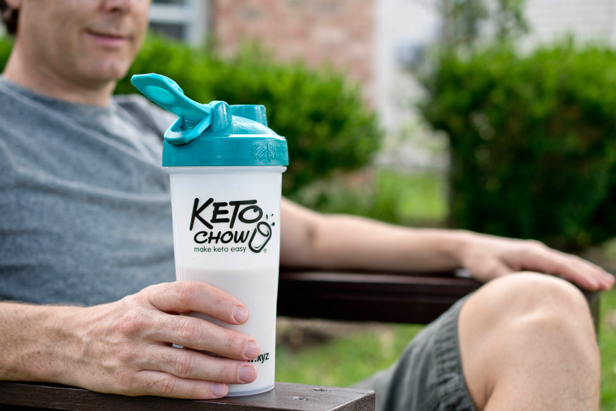 light colored keto chow in a teal blender bottle (most likely cookies and cream, banana, or vanilla) held by a man in a grey shirt.